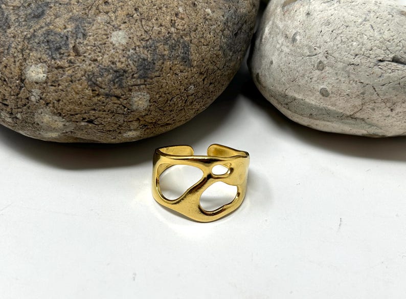 May include: A gold-colored adjustable ring with an open, abstract design. The ring features irregular cutouts, creating an artistic and modern look. The ring is displayed on a white surface, with two large, textured stones in the background.
