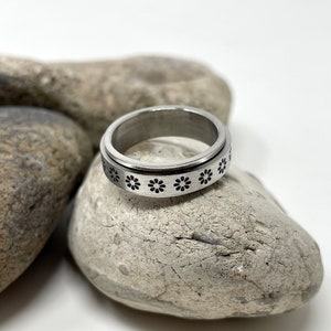 May include: A silver spinning ring with a black flower design. The ring is resting on a gray rock.