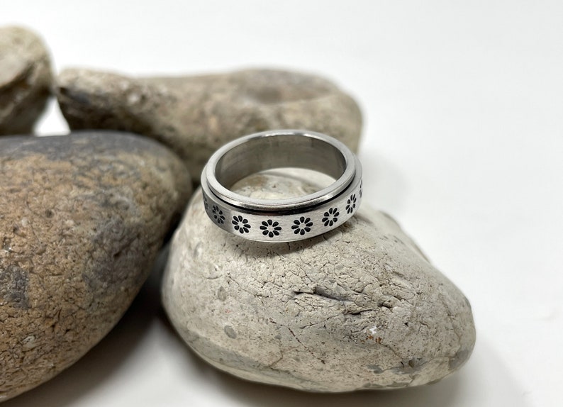 May include: A silver spinning ring with a black flower design. The ring is resting on a gray rock.