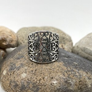 May include: A silver ring with a wide band featuring a delicate, intricate, cutout design of swirling floral patterns.