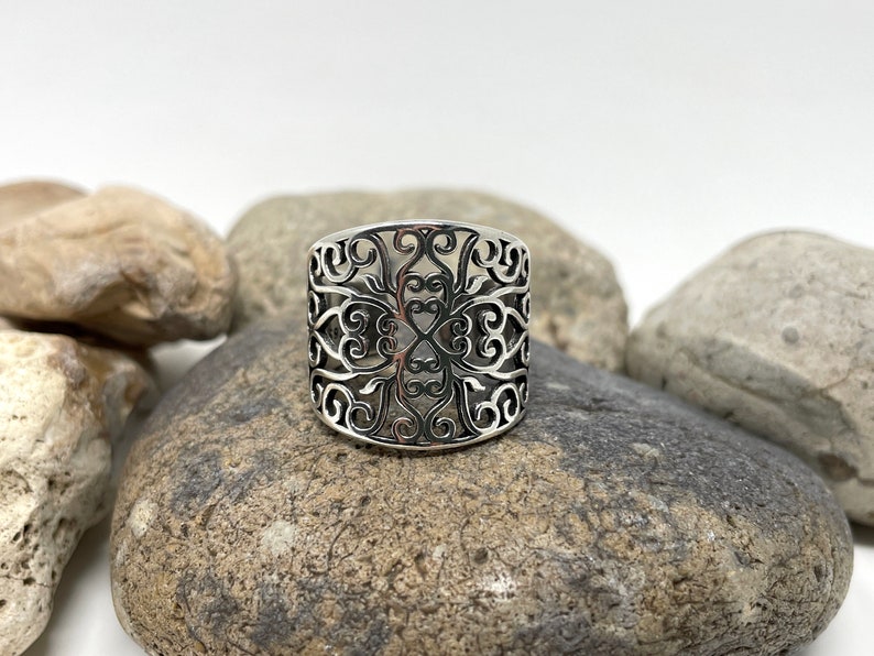 May include: A silver ring with a wide band featuring a delicate, intricate, cutout design of swirling floral patterns.