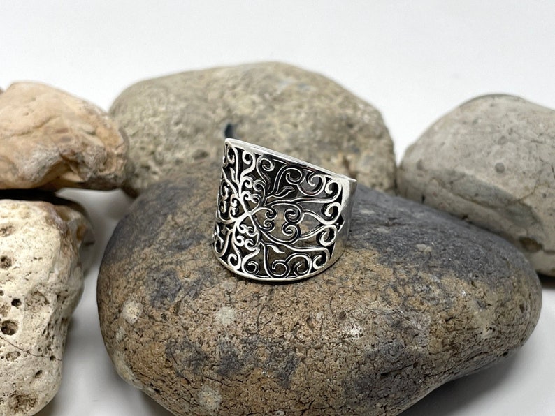 May include: A silver ring with a wide band featuring a delicate, intricate, cutout design of swirling floral patterns.