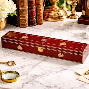 May include: A rectangular, deep red leather box with gold-toned trim and a central brass latch. The lid features three gold-embossed crown motifs and a central monogram. The box is positioned horizontally on a white marble surface.