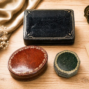 May include: Three small storage boxes: a rectangular black box, an oval reddish-brown box, and a hexagonal dark green box. Each box features a smooth, textured surface with intricate gold-coloured decorative filigree patterns along the edges.