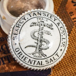 May include: A white ceramic jar with a lid featuring a black and white emblem. The emblem includes a snake wrapped around a tree, with text that reads "Oriental Salt". The jar sits on a patterned surface.