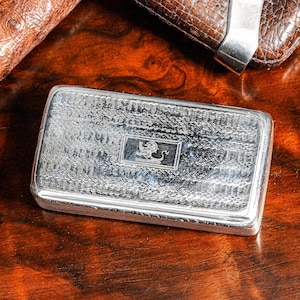May include: A rectangular silver-toned metal case featuring an intricate engine-turned pattern across the surface and a central rectangular emblem depicting a lion. The case has a polished finish and a hinged lid design with visible side detailing.