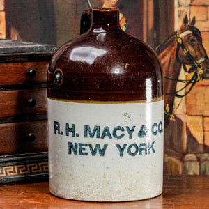 May include: A vintage stoneware jug with a brown glazed top and a cream-colored base. The jug features the text "R.H. MACY & CO NEW YORK" in blue lettering. The jug is displayed on a wooden surface.