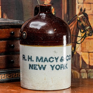 May include: Antique stoneware jug with a brown glazed top and a cream-colored base. The jug features the text "R.H. MACY & CO NEW YORK" in blue lettering. The jug is displayed on a wooden surface, with a painting in the background.