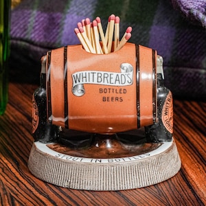 May include: A ceramic match holder shaped like a brown barrel with black bands, featuring the text Whitbread's Bottled Beers. The base is circular and textured with the text Stout In Bottles. A cluster of wooden matches with pink tips sits inside.