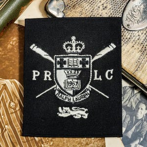 May include: A black square patch with a white crest design featuring crossed oars, a crown, and the text "PR LC" and "RALPH LAUREN 1967". The patch is surrounded by vintage photographs and a leather-bound book.