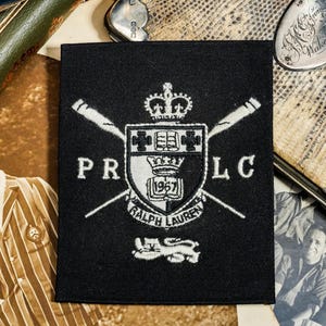 May include: A rectangular black fabric patch featuring white embroidered details. The design includes a central shield with a crown, two crossed oars, the letters PR and LC, the text Ralph Lauren, the year 1967, and a lion emblem at the bottom.