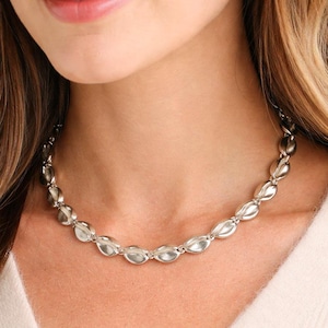 May include: A silver-toned necklace featuring a series of interconnected, smooth, oval-shaped links. Each link has a subtle, rounded, seed-like form, creating a continuous, polished chain that rests around the neck with a uniform, metallic luster.