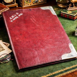 May include: A dark red leather-bound portfolio with silver-toned metal corner protectors. The cover features the embossed text A.M.L-S and 10 APRIL 1912. The portfolio is positioned on a green surface alongside vintage items including glasses.