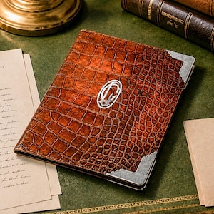 May include: A brown leather journal with a crocodile pattern, silver corner accents, and a silver emblem. The journal rests on a green desk next to a pen, inkwell, and vintage letters. Books are in the background.