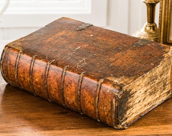 May include: A large, antique, rectangular book with a distressed, weathered brown leather cover featuring raised bands on the spine and worn edges. The book is positioned horizontally, showcasing its aged texture and dark, mottled brown finish.