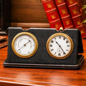 May include: A black leather-bound barometer and clock combination with gold-colored bezels. The barometer dial reads "Rain, Change, Fair" and the clock has Roman numerals. The items are on a wooden surface.