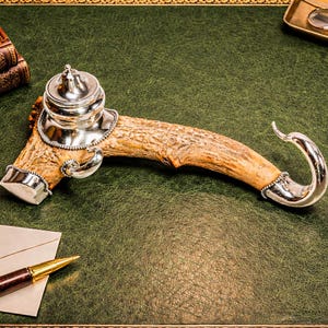 May include: An antique inkwell crafted from a deer antler, adorned with silver-plated accents. The inkwell sits on a green leather surface, accompanied by a pen, envelope, and vintage books. A magnifying glass and spectacles are also visible.