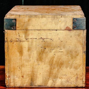 Puede incluir: Una caja de madera vintage de forma rectangular con tapa con bisagras. La caja est&aacute; pintada de color amarillo claro y muestra signos de desgaste, como ara&ntilde;azos y decoloraci&oacute;n. Las escuadras met&aacute;licas aseguran los bordes.