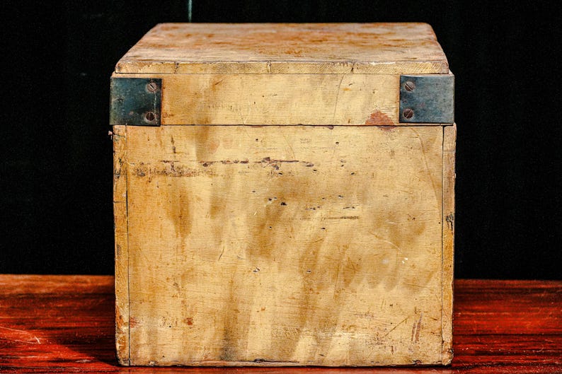 Puede incluir: Una caja de madera vintage de forma rectangular con tapa con bisagras. La caja est&aacute; pintada de color amarillo claro y muestra signos de desgaste, como ara&ntilde;azos y decoloraci&oacute;n. Las escuadras met&aacute;licas aseguran los bordes.