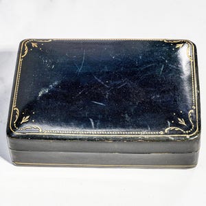 May include: A rectangular black box with a glossy finish and gold-coloured decorative filigree patterns along the edges. The lid features a thin gold border and corner accents, with visible surface scratches across the dark, reflective top surface.