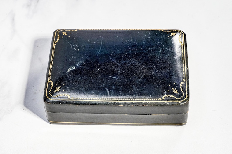 May include: A rectangular black box with a glossy finish and gold-coloured decorative filigree patterns along the edges. The lid features a thin gold border and corner accents, with visible surface scratches across the dark, reflective top surface.