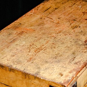Puede incluir: Primer plano de una caja de madera desgastada, con un tono marr&oacute;n dorado c&aacute;lido. La superficie muestra signos de desgaste, con ara&ntilde;azos y variaciones de color, lo que sugiere edad y uso. La construcci&oacute;n de la caja es visible en los bordes.