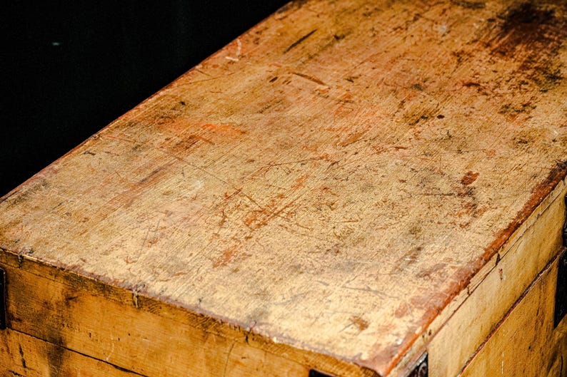 Puede incluir: Primer plano de una caja de madera desgastada, con un tono marr&oacute;n dorado c&aacute;lido. La superficie muestra signos de desgaste, con ara&ntilde;azos y variaciones de color, lo que sugiere edad y uso. La construcci&oacute;n de la caja es visible en los bordes.