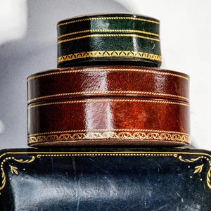 May include: A stack of three decorative boxes. The top box is small, round, and dark green with gold trim. The middle box is larger, round, and brown with gold detailing. The bottom box is rectangular, black, and features gold corner accents.