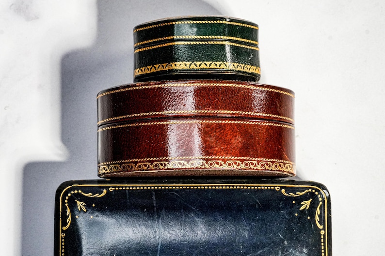 May include: A stack of three decorative boxes. The top box is small, round, and dark green with gold trim. The middle box is larger, round, and brown with gold detailing. The bottom box is rectangular, black, and features gold corner accents.
