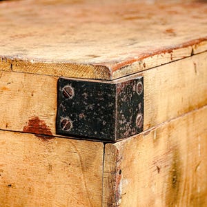 Puede incluir: Primer plano de un cofre de madera envejecida con esquinas met&aacute;licas y un pestillo. La madera tiene un tono dorado c&aacute;lido con vetas visibles y algunas manchas rojizas. La ferreter&iacute;a met&aacute;lica muestra signos de edad.