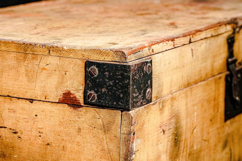 Puede incluir: Primer plano de un cofre de madera envejecida con esquinas met&aacute;licas y un pestillo. La madera tiene un tono dorado c&aacute;lido con vetas visibles y algunas manchas rojizas. La ferreter&iacute;a met&aacute;lica muestra signos de edad.
