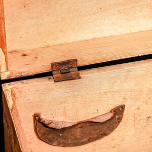 Puede incluir: Primer plano de una caja de madera abierta con una bisagra de metal oxidado y una pieza de metal curvada. La madera es de color marr&oacute;n claro con vetas visibles y algo de desgaste.