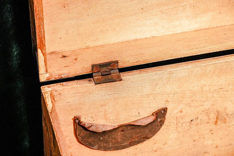 Puede incluir: Primer plano de una caja de madera abierta con una bisagra de metal oxidado y una pieza de metal curvada. La madera es de color marr&oacute;n claro con vetas visibles y algo de desgaste.