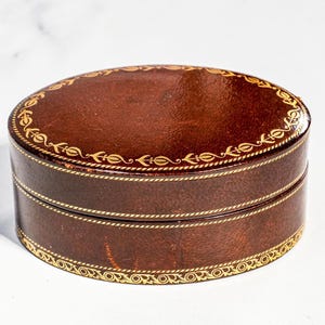 May include: An oval-shaped dark brown box with a lid, featuring gold-coloured decorative filigree patterns along the top edge and the base. The box has a smooth, polished finish and a consistent dark brown colour throughout its exterior surface.
