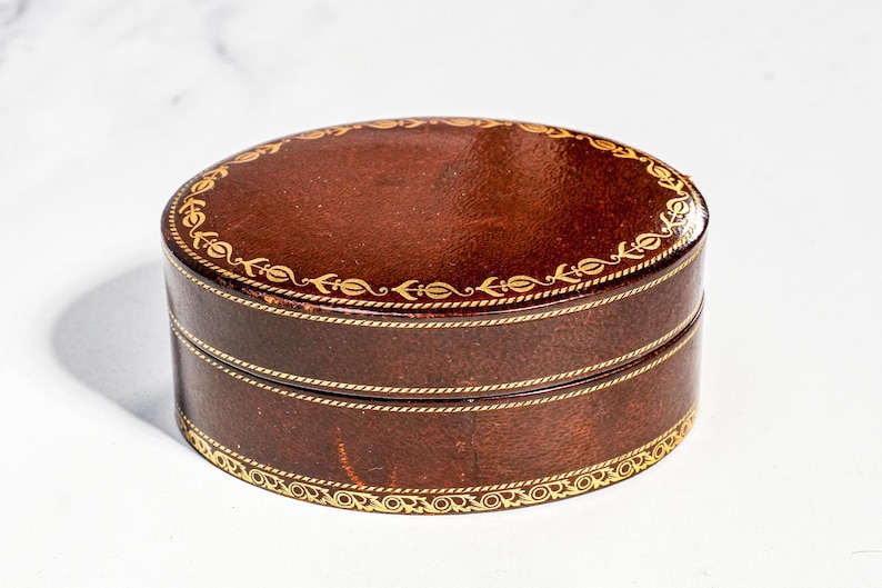 May include: An oval-shaped dark brown box with a lid, featuring gold-coloured decorative filigree patterns along the top edge and the base. The box has a smooth, polished finish and a consistent dark brown colour throughout its exterior surface.
