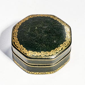 May include: An octagonal dark green box with a textured surface and gold-coloured decorative filigree patterns along the edges of the lid and the base. The box features a hinged lid and a horizontal seam running around the centre of the object.