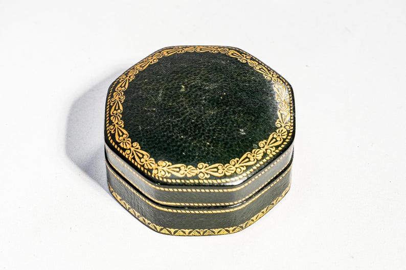 May include: An octagonal dark green box with a textured surface and gold-coloured decorative filigree patterns along the edges of the lid and the base. The box features a hinged lid and a horizontal seam running around the centre of the object.