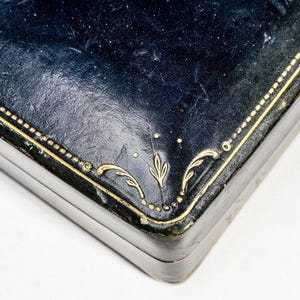 May include: A rectangular dark blue box with a textured surface featuring gold-coloured decorative lines and leaf-like patterns embossed along the edges. The lid has a smooth, slightly worn finish with visible gold detailing at the corner.