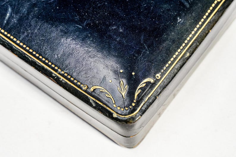 May include: A rectangular dark blue box with a textured surface featuring gold-coloured decorative lines and leaf-like patterns embossed along the edges. The lid has a smooth, slightly worn finish with visible gold detailing at the corner.