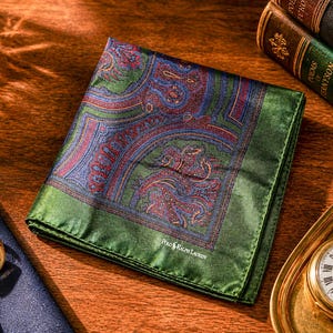 May include: A green and paisley patterned pocket square with red and blue accents. The pocket square is next to a navy blue tie with a gold button, a gold pocket watch, and two antique books.