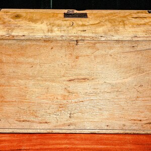 Puede incluir: Un cofre de madera rectangular con un acabado marr&oacute;n claro. El cofre tiene una tapa con bisagras y herrajes met&aacute;licos en las esquinas y el cierre. La veta de la madera es visible y el cofre parece envejecido.
