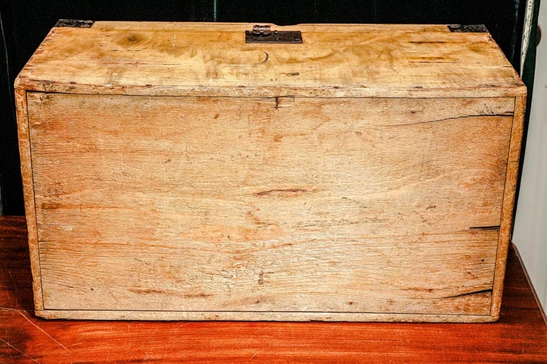 Puede incluir: Un cofre de madera rectangular con un acabado marr&oacute;n claro. El cofre tiene una tapa con bisagras y herrajes met&aacute;licos en las esquinas y el cierre. La veta de la madera es visible y el cofre parece envejecido.