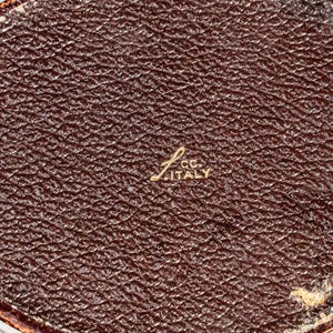 May include: An oval-shaped object with a dark brown, textured surface resembling leather. In the centre, gold-coloured text is embossed, reading "L. CC. ITALY". The edges of the object show a thin, worn, reddish-brown border with visible wear.