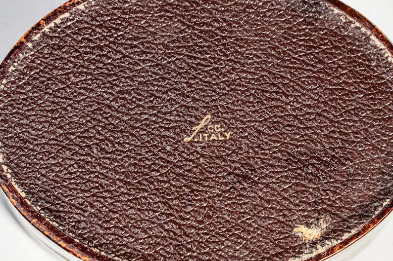 May include: An oval-shaped object with a dark brown, textured surface resembling leather. In the centre, gold-coloured text is embossed, reading "L. CC. ITALY". The edges of the object show a thin, worn, reddish-brown border with visible wear.