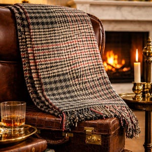 May include: A cozy, patterned throw blanket draped over a leather chair. The blanket features a black, gray, and red houndstooth design with fringe. A leather suitcase and a glass of amber liquid sit nearby, suggesting a warm, inviting atmosphere.