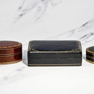 May include: Three decorative boxes: a brown oval box, a rectangular black box, and a small hexagonal black box. Each box features a smooth finish and intricate gold-coloured filigree detailing along the edges and lids, providing a uniform look.