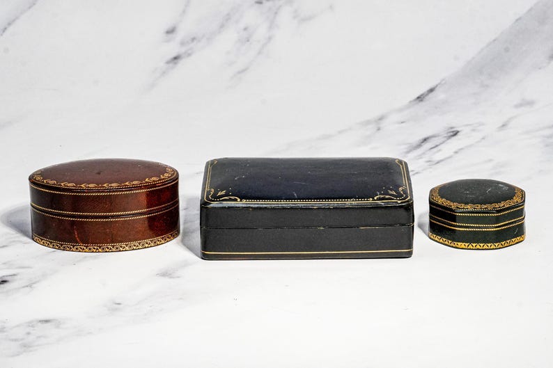 May include: Three decorative boxes: a brown oval box, a rectangular black box, and a small hexagonal black box. Each box features a smooth finish and intricate gold-coloured filigree detailing along the edges and lids, providing a uniform look.