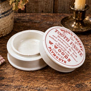 May include: A white ceramic jar with a matching lid, featuring red printed text that reads Woods Cherry Tooth Paste, For removing Tartar and whitening the Teeth without injuring the Enamel, W. Woods Chemist Plymouth, and 6d.