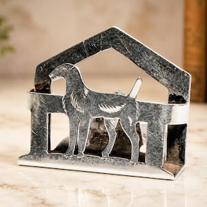May include: A silver-toned metal napkin holder featuring a cutout silhouette of a standing dog centered within a house-shaped frame. The metal surface has a distressed, textured finish with visible dark markings throughout the structure.