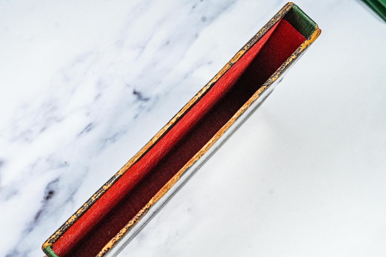 Puede incluir: Una caja rectangular con interior rojo y exterior oscuro con ribete dorado. La caja est&aacute; abierta, revelando el forro rojo. Los bordes son verdes. La caja est&aacute; sobre un fondo de m&aacute;rmol blanco.